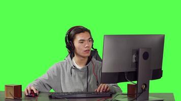 Young player sitting at computer desk and playing online video games with friends, using modern devices to have a good time. Asian man participating in rpg tournament over greenscreen.