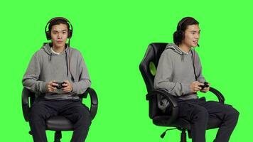 Confident guy playing video games in studio, having fun with friends on online gaming quest over greenscreen backdrop. Asian man gamer enjoying rpg competition sitting on chair.