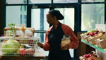 Afrikaanse Amerikaans vrouw in lokaal supermarkt bijvullen schappen met vers geoogst produceren, klein bedrijf eigenaar in buurt. winkelier regelen van bijkeuken producten in herbruikbaar containers. video