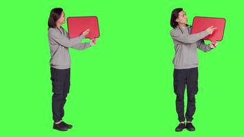 Male model poses with speech bubble, presenting blank cardboard with copyspace against full body greenscreen. Asian person holding red carton board for marketing advertisement. video