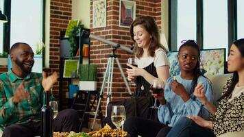 Best friends having fun together, enjoying alcohol beverages and eating crackers in cozy apartment living room. BIPOC group of people chatting at home, gathered together for anniversary video