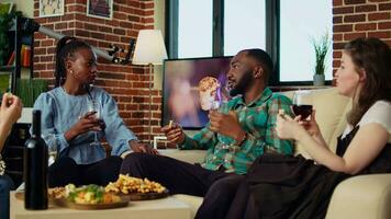 mångkulturell grupp vänner fira afrikansk amerikan man födelsedag, dricka vin och äter snacks i lägenhet. gäster skrattande och talande på hus sammankomst efter erbjudande värd presenterar video