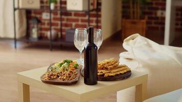 Panning shot of appetizers board and gourmet cheese platter set up by hosts to properly welcome guests arriving soon at social gathering. Wine bottle and glasses next to food in cozy home, close up video