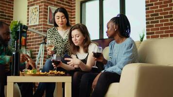 Multiethnic friends at reunion looking at funny videos from their childhood on smartphone screen. Cheerful guests in living room watching clips from their youth on mobile phone