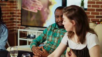 afrikanisch amerikanisch Mann chatten mit Freundinnen auf Couch beim Wohnung Party, genießen Gespräch. gemischtrassig Paar eingeladen beim freunde Geburtstag, reden und Trinken Alkohol video