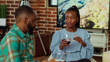 africano americano hombre a social reunión hablando con su Novia en sofá mientras disfrutando vaso de vino. marido y esposa invitados a Departamento fiesta teniendo agradable conversacion en ladrillo pared hogar video