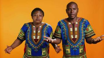 Clueless man and woman doing i dont know sign in studio, expressing confusion and being uncertain about answer. African american people in doubt being unsure over orange background video