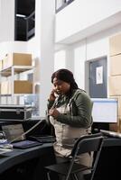 Employee using landline phone to talk with remote client, discussing order detalis before start preparing package for delivery. African american worker wearing industrial overall working in storehouse photo