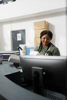 Stockroom worker looking at white cardboard boxes, checking customers orders on computer in storage room. African american employee wearing industrial overall while preparing packages for shipping photo