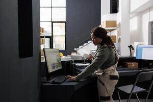 Warehouse employee looking at carton white boxes, checking shipping details on computer in storage room. African american supervisor preparing customers orders working at goods quality control photo