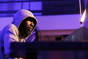 African american internet thief planning phishing attack and online scam in hideout workspace. Hacker coding computer virus and ransomware to break into server and steal data photo