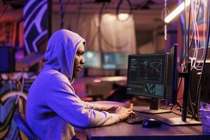 African american hacker coding spyware in abandoned warehouse. Man in hood using computer for breaching data, cracking password and getting access to server database at night photo
