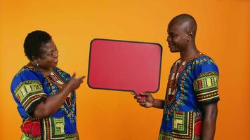 Ethnic young couple presenting speech bubble board, creating marketing advertisement and promoting red billboard empty sign in studio. African american people pointing to cardboard icon. video