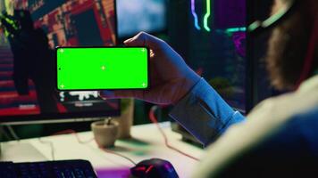 Man in dimly lit apartment watching tutorial on how to finish videogame on green screen smartphone, enjoying day off. Gamer learning online multiplayer shooter tips from video on mockup mobile phone