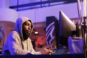 African american criminal hacking online server and stealing information. Hacker in hood planning phishing attack and programming malicious software in abandoned warehouse photo