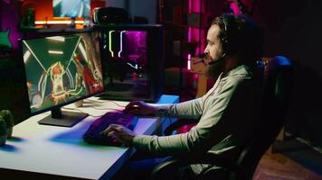 Happy man using headphones mic to communicate with online friends from his dark living room, playing engaging video games on gaming PC at computer desk, relaxing after work