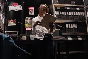 Woman detective analyzing information from forensic expertise conclusion while standing in private agency room. African american police investigation reading crime case file at night photo