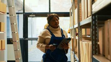 African american woman scanning barcodes on boxes, working with scanner and tablet to check merchandise o shelves and racks. Female employee looking at retail products for depot inventory. photo