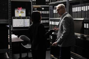 Accountancy executives discussing quarterly profit revenue, analysing performance data. Asian woman and senior businessman in bureaucratic workplace surrounded by documentation folders in cabinets photo