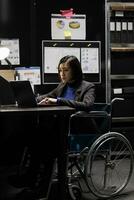 paralizado mujer de negocios contabilidad ejecutivo en silla de ruedas agregando presupuesto plan datos en ordenador portátil. asiático mujer con paraplejía en almacenamiento oficina lleno con papeleo carpetas en estantería y diagramas de flujo foto