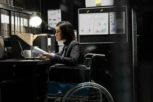 Paralyzed businesswoman employee in wheelchair checking bookkeeping analytical data documents. Accountant with disability in business company file cabinet depository filled with charts and folders photo