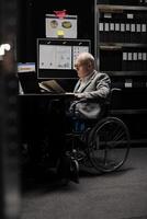 Senior wheelchair user checking annual profit revenue paperwork before business consulting meeting. Executive with paraplegia doing objective completion measurements in file cabinet office photo