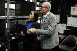 Teamworking executives coworkers in storage repository office looking for accounting bureaucratic paperwork on file cabinet shelves. Elderly businessman and asian woman in archival room photo