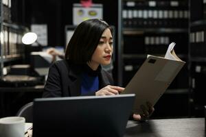Asian businesswoman executive checking accountancy budget plan data paperwork. Bureaucratic administration employee in repository filled with document folders and graphical chart reports photo