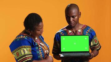 Ethnic couple pointing at laptop with greenscreen display, posing in studio with portable pc presenting isolated copyspace layout. Man and woman looking at blank mockup template on camera. video