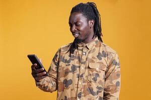 African american man reading job application on smartphone, scrolling through social media in studio over yellow background. Cheerful young adult searching information on mobile phone photo