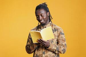 alegre africano americano hombre leyendo ficción libro, disfrutando aprendizaje nuevo información en pie en estudio terminado amarillo antecedentes. inteligente persona estudiando literatura, gusto género y trama foto