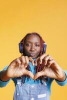 Person doing heart shape sign in studio, listening to music on headphones. Female model showing romantic symbol and love gesture on orange background, advertising romance and flirt. photo