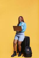 Young person using laptop in studio over orange background, browsing internet website on camera. Female model with trolley bags holding wireless pc to check social media page. photo