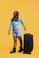 Smiling woman listening to music on flight, having big luggage trolley for weekend getaway or city break. Happy young adult wearing headset on vacation journey, posing in studio. photo