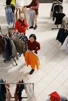 Diverse people shopping for clothes in retail store, looking for christmas presents and festive attire at mall. Women checking fabric of clothing items on hangers, buying trendy merchandise. photo