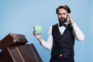Sick worker displaying package of drugs against blue background while struggling with headache and using medication for pain relief. Concierge at hotel takes analgesics as he feels ill. photo
