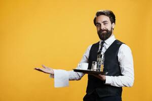 Professional server presenting something in studio, providing luxurious service at restaurant while he carries platter with cutlery and coffee. Male butler serving drinks to clients, fine dining. photo