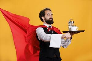 Strong waiter serves food on tray while he is wearing formal suit and hero mantle, superhuman concept at luxury restaurant. Young man butler carry platter with eatery, professional dining. photo