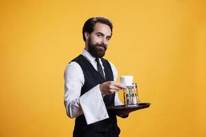 experimentado camarero servicio café a mesa, fingiendo a mano fuera taza de refresco para clientes. joven adulto mayordomo trabajando a lujo restaurante, que lleva comida bandeja para abastecimiento. foto