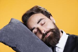 Tired waiter falling asleep on camera, taking a nap while he in standing against yellow background. Young sleepy person being overworked with burnout, falling asleep on pillow in studio. photo