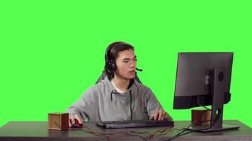 Young gamer sitting at computer desk and playing web based video games with people, using modern gadgets to have a good time. Asian person competing in rpg contest over greenscreen.