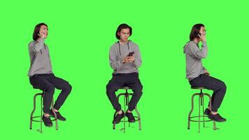 Asian guy texting and answering call on greenscreen backdrop, sending messages on social media using modern phone app. Young adult sitting on chair, calling family and messaging. video