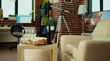 Panning shot of empty cozy apartment living room with appetizer platter and wine bottle on wooden table, awaiting guests to arrive. House interior with leather couch and TV running in background video