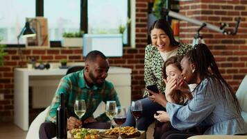 multiracial copains à appartement fête à la recherche à social médias des postes fabriqué par connaissance sur téléphone portable, fabrication amusement de eux. amusé invités dans vivant pièce réagir à reculer images publié en ligne par camarade video