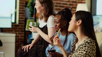 bipoc Dames zittend Aan bankstel Bij huis, roddelen over hun vriendjes terwijl genieten van glas van wijn en snacks. multi-etnisch vrienden drinken alcohol en chatten in steen muur huis video