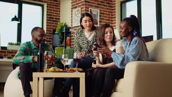 Friends at house party using smartphone to laugh at social media posts made by acquaintance while eating snacks and drinking wine. Guests using phone to look at images online, enjoying appetizers video