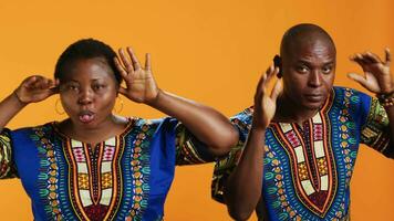 Silly ethnic couple fooling around on camera with comic face, having fun and sticking tongue out in studio. Cheerful people doing funky gesture and enjoying time on set, orange background. video