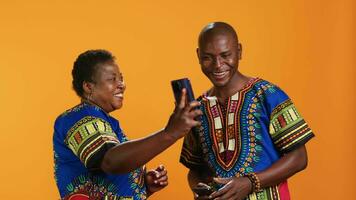 Traditional african american couple browsing websites on smartphone, checking photos on social media app and laughing at memes. Joyful people in colorful attire looking at pictures online. video