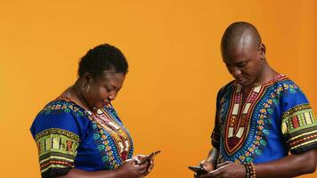 glücklich afrikanisch amerikanisch Menschen Scrollen Webseiten auf Smartphone, Überprüfung Bilder auf Sozial Medien App und haben Spaß mit Meme. verheiratet Paar im traditionell Kleidung suchen beim Fotos online. video