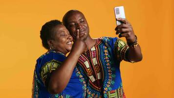 Smiling ethnic couple taking photos with phone in studio, having fun making memories with smartphone camera. Cheerful people take pictures while they wear traditional clothing. video